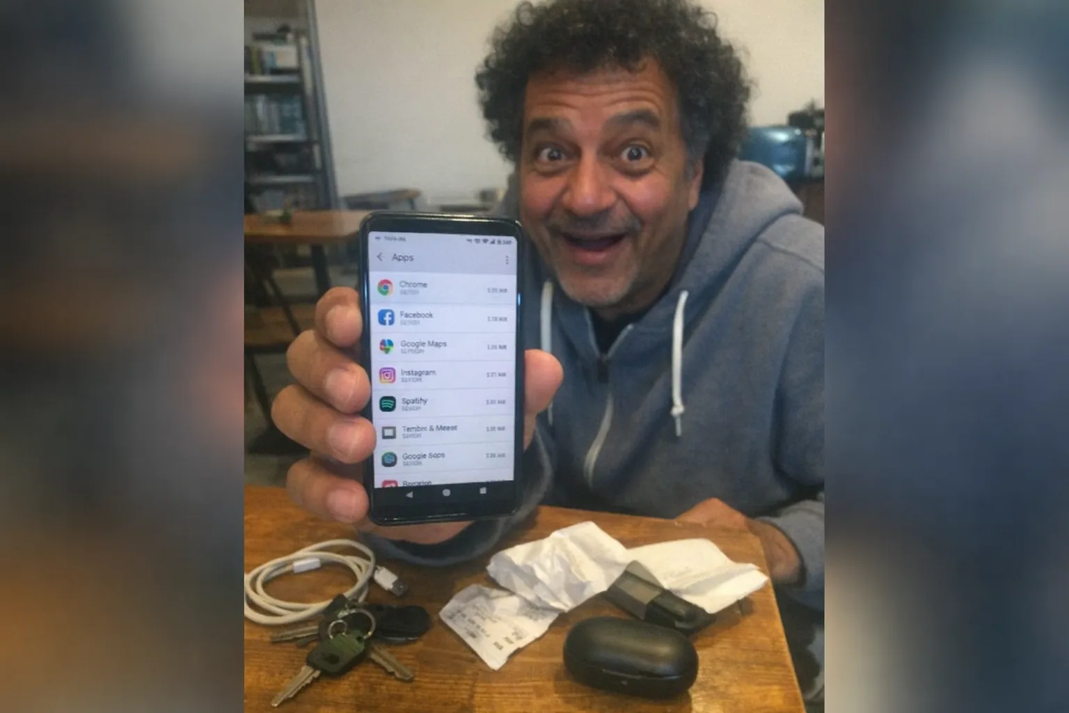Realistic iPhone snapshot in a small cafe: a mid-50s North African man with warm brown skin, average build, medium-length curly hair with natural volume, wearing streetwear (hoodie and sneakers), holding an Android phone close to the camera with the Settings app open to an apps list; his expression is amused and surprised (small grin with eyes crinkling, wide eyes, slightly open mouth) and his posture looks awkward/uncertain. On the table: a messy spread of everyday items (keys, charging cable, crumpled receipt, generic wireless earbuds case, and a small unbranded gadget partially tucked under a napkin). Aggressively mediocre composition, awkward framing, slight motion blur, mildly overexposed uneven lighting, visible noise/grain, candid phone photo vibe. No text overlays, no logos, no brand names, non-explicit.