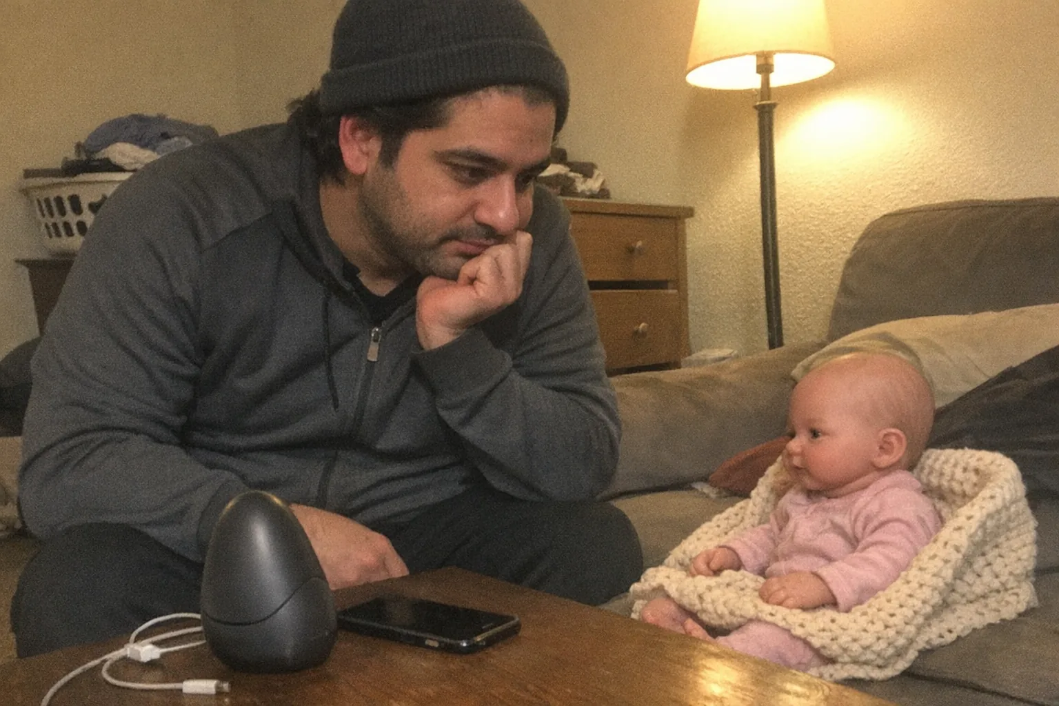 A candid iPhone snapshot in a small, slightly cluttered apartment living room at dusk, awkward framing and aggressively mediocre composition, slight motion blur and uneven warm lamp lighting with visible noise/grain. Primary subject: a calm, thoughtful Middle Eastern man in his early 30s with olive skin, dad-bod build, medium-length hair under a beanie, wearing athleisure (zip-up jacket and joggers). He’s leaning in with curious focused eyes and one hand on his chin, looking at a hyper-realistic reborn baby doll sitting on a couch with a tiny knitted blanket. On a coffee table nearby is a sleek, unbranded piece of adult-tech hardware (non-explicit, ambiguous shape) next to a phone and a charging cable, suggesting modern companionship tech without showing any branding or text. Background includes everyday mess: laundry basket, half-open drawer, a cheap floor lamp; mildly overexposed highlights from the lamp, no captions, no logos, no watermarks.