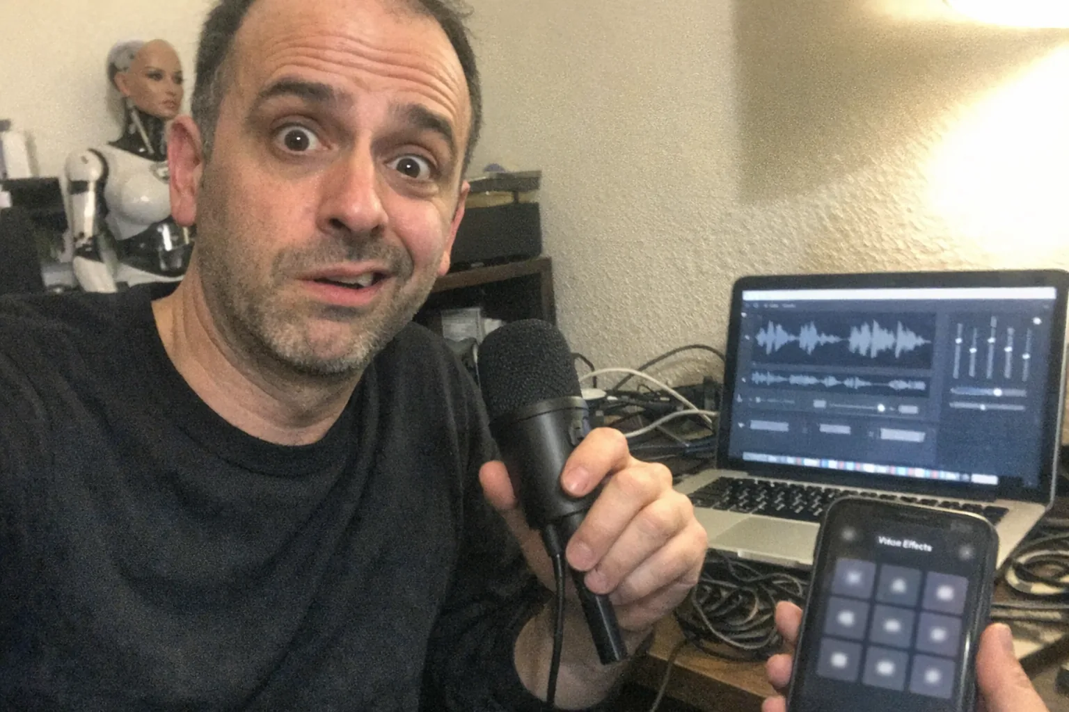 Realistic iPhone snapshot, aggressively mediocre composition: a slim white man in his early 40s with an olive skin tone, thinning short hairline, wearing a minimalist monochrome outfit (plain black crewneck and dark pants), slouched posture with faint under-eye circles, making a surprised and skeptical expression (wide eyes, slightly open mouth, one raised eyebrow with a half-smirk) while holding a cheap USB microphone near his laptop. On the laptop screen there are generic audio waveforms and sliders (no readable text). A small, non-explicit feminine humanoid robot bust sits on a cluttered shelf in the background, slightly out of focus. Casual home office setting with messy cables, a headset on the desk, and a phone showing an unlabeled voice effects grid (no logos). Awkward framing, slight motion blur, mildly overexposed uneven lamp lighting, visible noise/grain, candid unpolished feel, no text, no watermarks, no brand names.