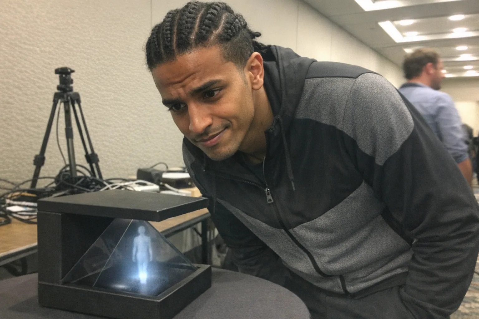 Realistic iPhone snapshot in a conference hallway demo area: a slim North African man in his mid-20s with warm brown skin, coily hair in neat cornrows/braids, wearing athleisure (zip-up jacket and joggers). He’s leaning toward a small tabletop “hologram” display box showing a faint floating human figure silhouette (non-explicit), looking determined (set jaw, intent gaze) but also awkward (uncertain posture, hesitant smile). Background includes a cluttered folding table with cables, a cheap tripod, and a second person out of focus who does NOT match the same appearance. Awkward framing, slight motion blur, mildly overexposed overhead fluorescent lighting, uneven white balance, natural phone noise/grain, aggressively mediocre composition. No text, no captions, no logos, no watermarks.