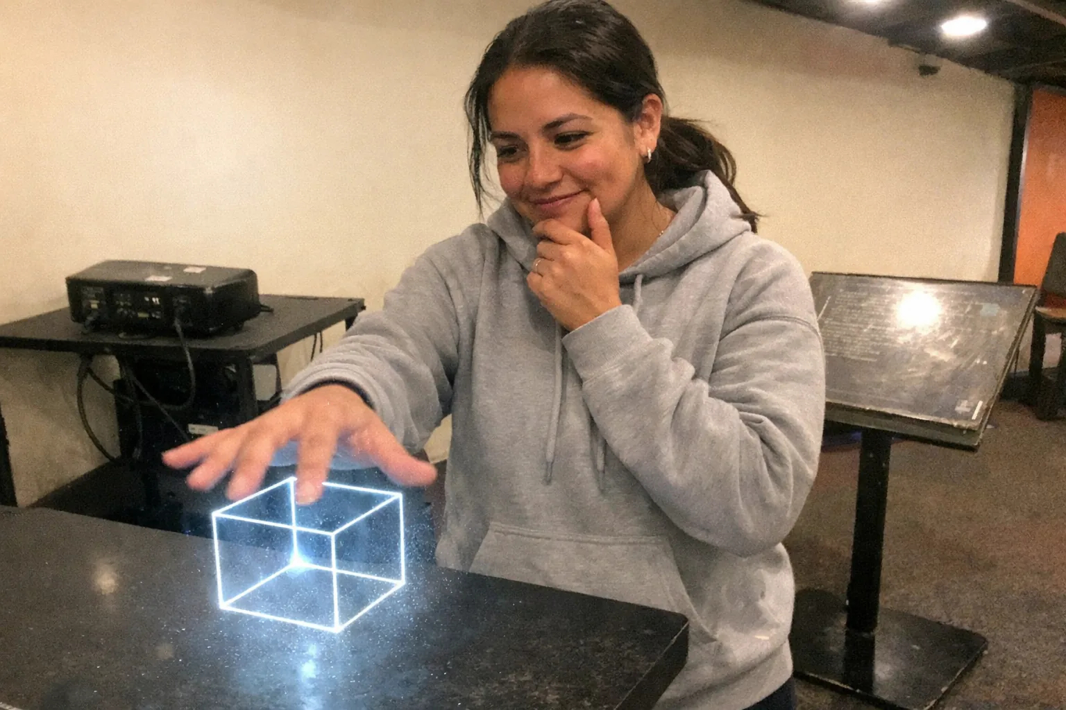 A candid iPhone snapshot in a small science museum exhibit area: a Latina woman in her early 30s with tan skin and a muscular (not bodybuilder) build stands slightly off-center, awkwardly framed from mid-thigh up. She wears streetwear (a plain hoodie and sneakers), hair in a low ponytail with a few loose strands. Expression cues: amused small grin with eyes slightly crinkling, thoughtful hand on chin while her other hand reaches toward a faint “hologram-like” projection hovering above a table (a simple light projection with visible dust/mist in the beam). The projection looks like a floating wireframe cube, and her hand is visibly passing through it. Mild motion blur on the reaching hand, slightly overexposed overhead lighting, uneven color temperature, natural phone noise/grain, aggressively mediocre composition. Background includes mundane museum fixtures: a cheap black projector, cables, and a worn informational stand (but no readable text). No logos, no watermarks, no captions.