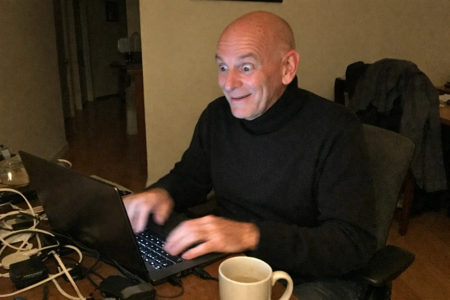 A candid iPhone snapshot in a cluttered home office at night: a slim White man in his early 60s with fair skin and a clean shaved head, wearing a minimalist monochrome outfit (plain black turtleneck and dark pants), seated at a small desk with a laptop open (screen glow visible but no readable text). He looks surprised and amused at the screen (wide eyes, slightly open mouth, small grin) but with a calm, steady posture. On the desk are messy cables, a generic unbranded small gadget, and a coffee mug; in the background a dim hallway and a coat on a chair. Awkward framing, slightly motion-blurred hands, mildly overexposed laptop glow, uneven warm lighting, natural phone noise/grain, aggressively mediocre composition. No text, no logos, no brand names, modern realistic scene.