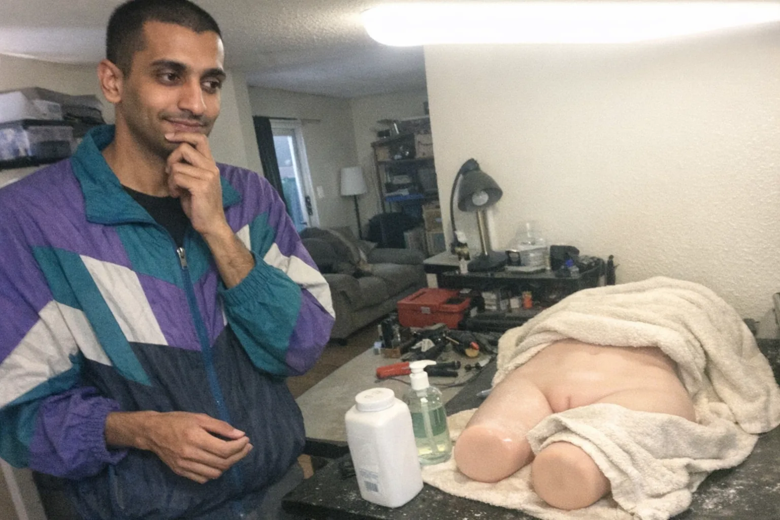 Realistic iPhone snapshot in a small apartment home-office/workbench area: a tall, lanky South Asian man in his early 30s with brown skin, dark buzz cut, wearing a retro vintage windbreaker, standing beside a cluttered table with a silicone doll limb/torso (non-explicit, partially covered by a plain towel) and a small container of body powder and mild soap. He looks thoughtful with one hand on his chin, calm soft expression, but slightly nervous with fidgety other hand and a tight smile. Awkward framing, slightly motion-blurred, mildly overexposed overhead light, uneven shadows, natural phone noise/grain, aggressively mediocre composition. No text, no logos, no watermarks; plausible candid maintenance moment.