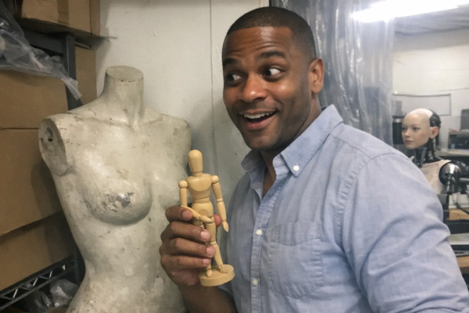 A realistic iPhone snapshot in a cluttered retail backroom/workshop: a male in his late 30s with medium-brown Black skin tone, athletic build with broad shoulders, dark buzz cut, wearing a business-casual button-down shirt with rolled sleeves. He’s holding a small wooden artist manikin in one hand while glancing toward a scuffed store mannequin torso leaning against a wall; his expression is amused and surprised (small grin, eyes crinkling, wide eyes, slightly open mouth). In the background, partially visible behind a plastic curtain, there’s a female humanoid robot prototype head-and-shoulders on a workbench (non-explicit, just a realistic robotics prop). Awkward framing, slight motion blur, mildly overexposed fluorescent lighting, natural phone noise/grain, aggressively mediocre composition, candid unremarkable moment. No text, no logos, no watermarks.