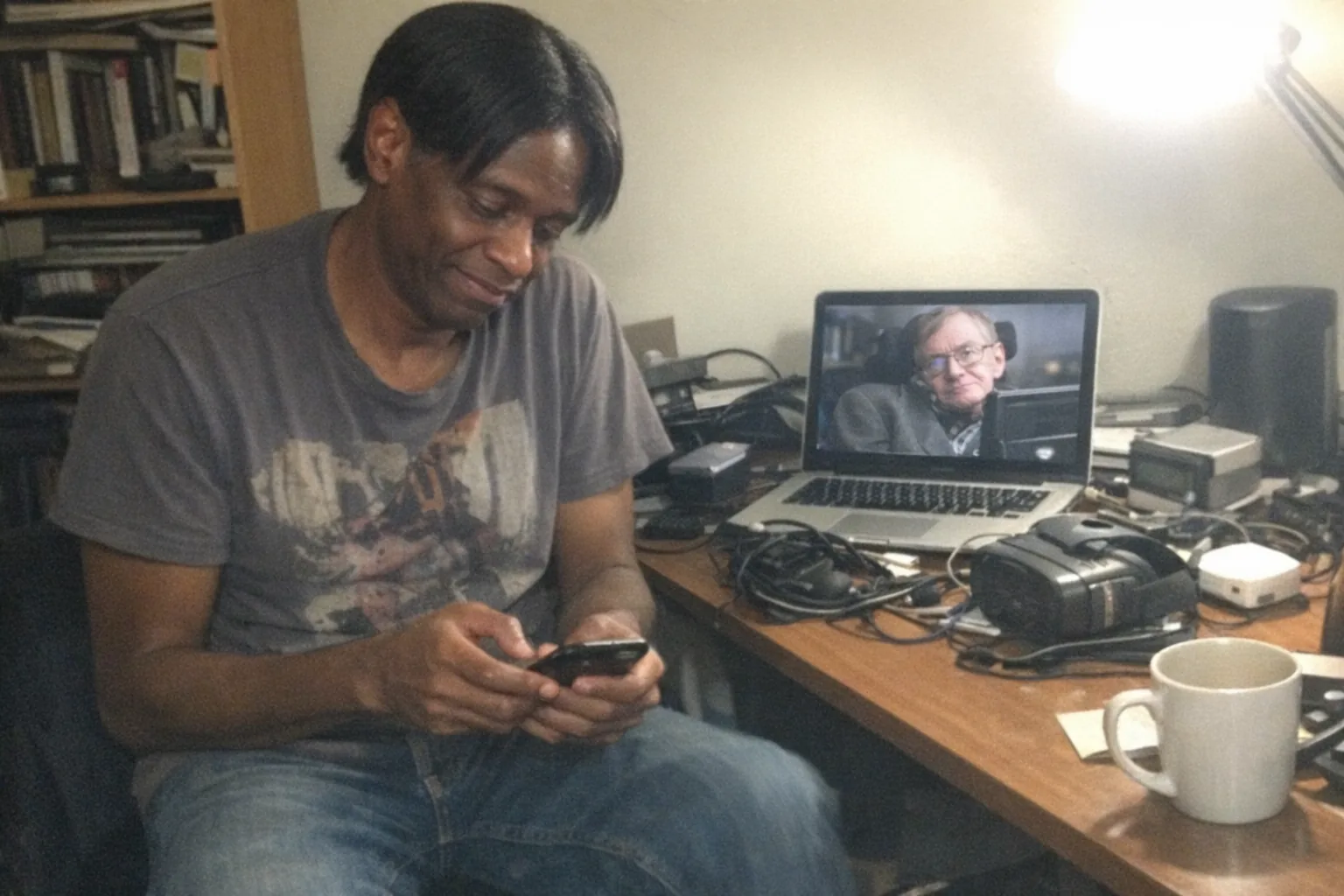 Realistic iPhone snapshot in a cramped home office at night, aggressively mediocre composition with awkward framing, slight motion blur, mild overexposure from a desk lamp, visible phone noise/grain. Primary human: a tall, lanky Black man (late 40s, dark brown skin) with straight hair neatly side-parted, wearing a casual graphic tee and jeans; he looks impatient (foot tapping), tired (slouched posture, faint under-eye circles), and nervous (fidgety hands, tight smile) while checking his phone. On his laptop screen (not readable text), a paused documentary frame of Stephen Hawking is visible, with a cluttered desk of cables, an old VR headset, and a small generic prototype gadget beside it (non-explicit). Background includes a messy bookshelf and a half-empty coffee mug; no logos, no brand names, no captions, no watermarks.