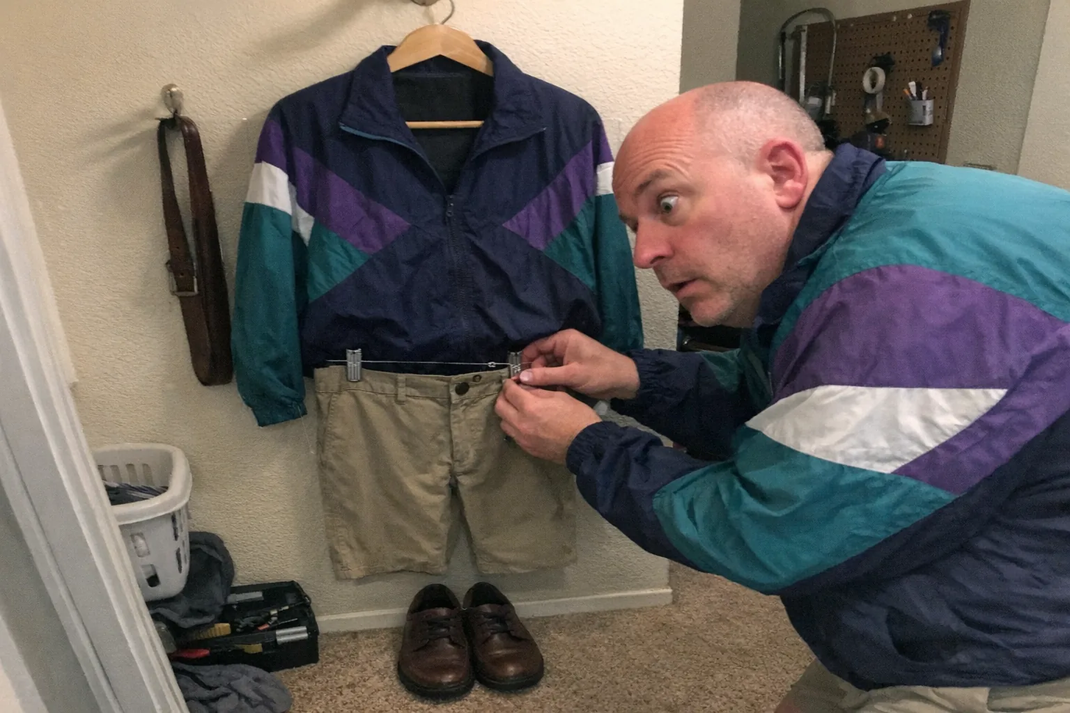 Realistic iPhone snapshot, candid and slightly awkward framing, mild motion blur and uneven indoor lighting, visible natural noise/grain, aggressively mediocre composition. Scene: a cluttered apartment hallway corner with a wall hook and a “floating” outfit display—shirt on a wide hanger, pants clipped below, shoes on the floor, a belt draped neatly on a side hook; fishing line and small clips are barely visible. Primary human: a white man in his early 50s with fair skin, dad-bod build, clean shaved head, wearing a retro vintage windbreaker; he’s leaning in close with focused eyes and a surprised expression (wide eyes, slightly open mouth) as he adjusts a clip on the waistband. Background includes everyday mess: a laundry basket, a small tool kit, and a pegboard with random hooks. No text, no captions, no watermarks, no logos, no brand names.