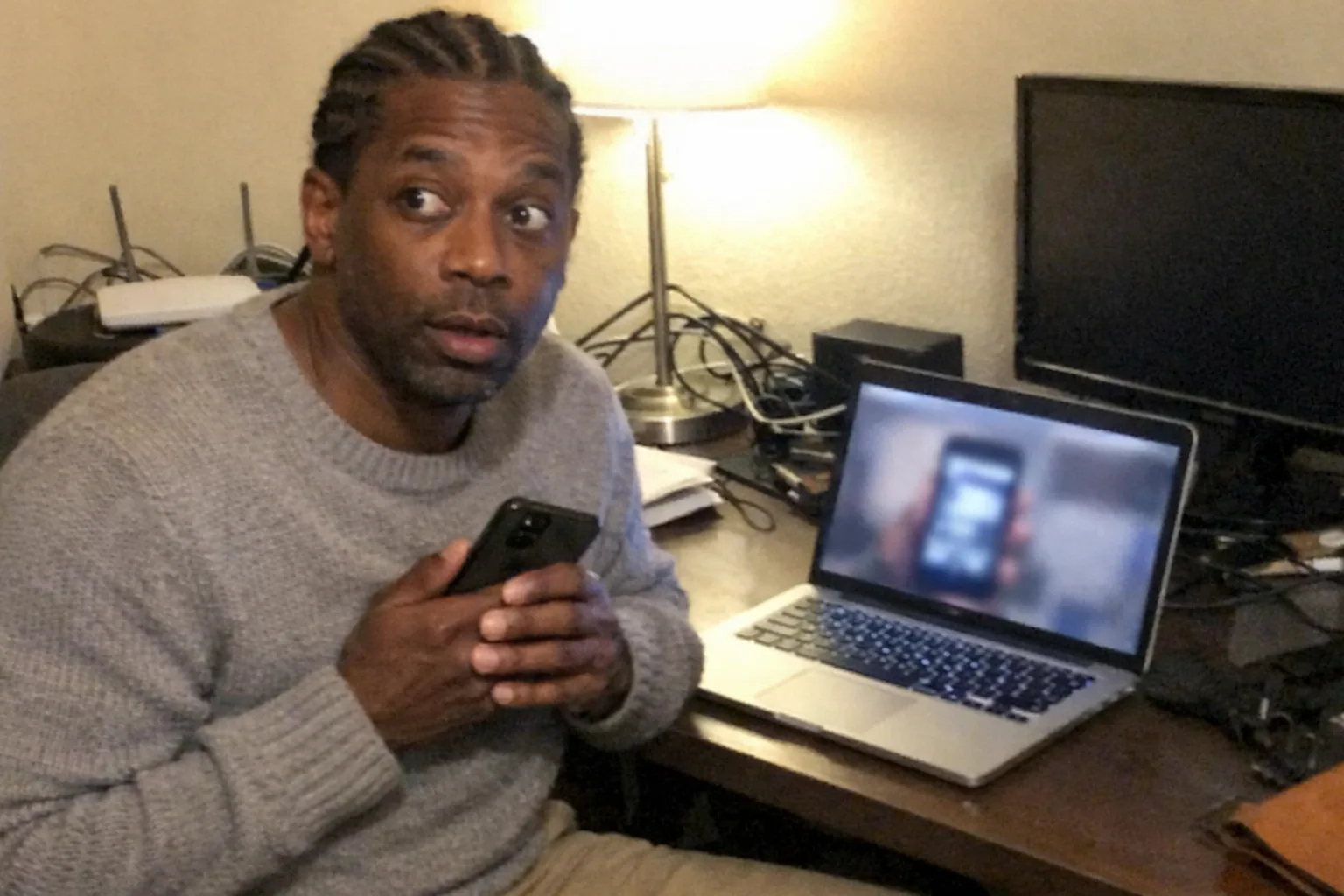 A realistic, aggressively mediocre iPhone snapshot taken in a cluttered home office at night, awkward framing and slight motion blur, mildly overexposed lamp lighting with natural phone noise/grain. Primary subject: a Black man in his early 40s with dark brown skin, lean runner build, coily hair in neat braids/cornrows, wearing a smart-casual knit sweater and chinos. His expression shows surprised wide eyes and slightly open mouth, but also tired slouched posture with faint under-eye circles and a calm soft steadiness. He’s holding a smartphone close to his chest while glancing at a laptop on the desk that appears to be showing a blurry mirrored version of the phone screen (generic shapes, no readable text). Background includes messy cables, a router, and a second monitor turned off. No logos, no brand names, no on-screen text, candid unpolished look.