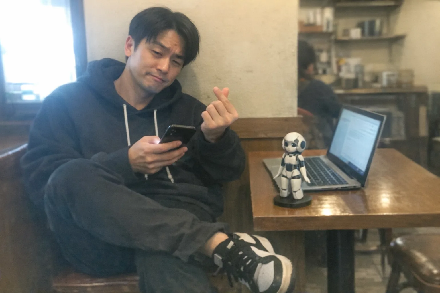 A casual iPhone snapshot in a small coffee shop, awkward framing and aggressively mediocre composition, slight motion blur and mild overexposure from a bright window. Primary subject: an East Asian man in his early 30s with light skin, slim build, straight neat side-parted hair, wearing streetwear (hoodie and sneakers), slouched posture with faint under-eye circles, tapping his foot while checking his phone, making a small finger-heart gesture with one hand; expression shows a skeptical raised eyebrow and half-smirk, like he’s unsure if the emoji was flirty. On the table: a laptop open (no readable text), a phone screen glow, and a small unbranded humanoid female robot figurine or compact assistant device nearby (non-explicit, just techy). Uneven indoor lighting, natural noise/grain, candid everyday vibe, no logos, no watermarks, no text.